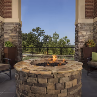 Stone fire pit with a small fire burning, surrounded by chairs and large stone pillars, overlooking a scenic view of trees and a clear blue sky.