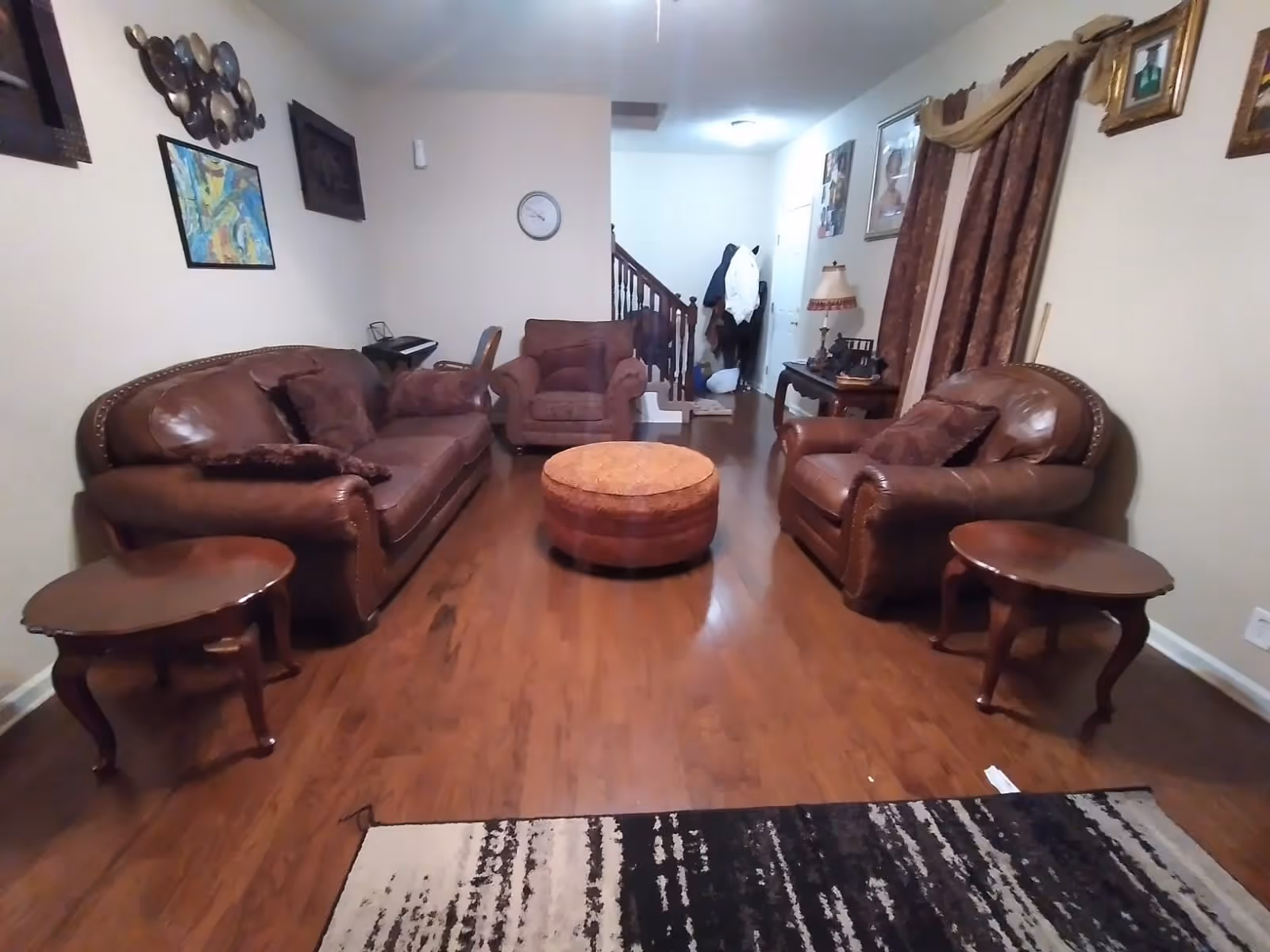 A living room with brown leather sofas and armchairs arranged around a round ottoman. There are two wooden side tables, a patterned rug on the wooden floor, and various framed pictures and decorations on the walls. A staircase and a coat rack are visible in the background.