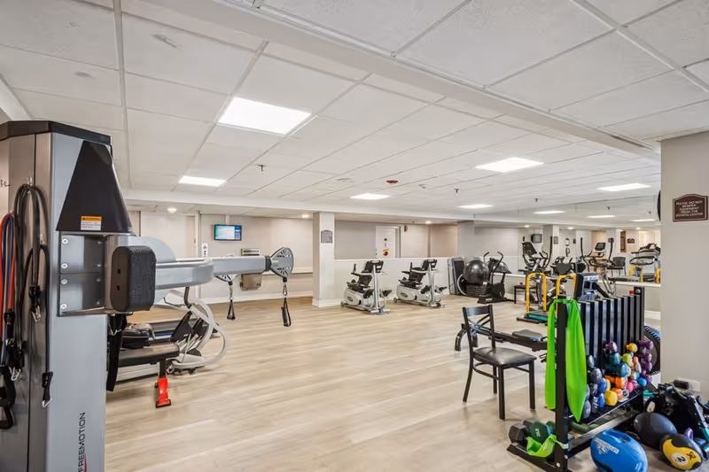 A spacious fitness room with various exercise equipment including stationary bikes, weight machines, dumbbells, resistance bands, and exercise balls. The room has light wood flooring, white walls, and a ceiling with recessed lighting. A large mirror covers one wall, reflecting the equipment and enhancing the sense of space.