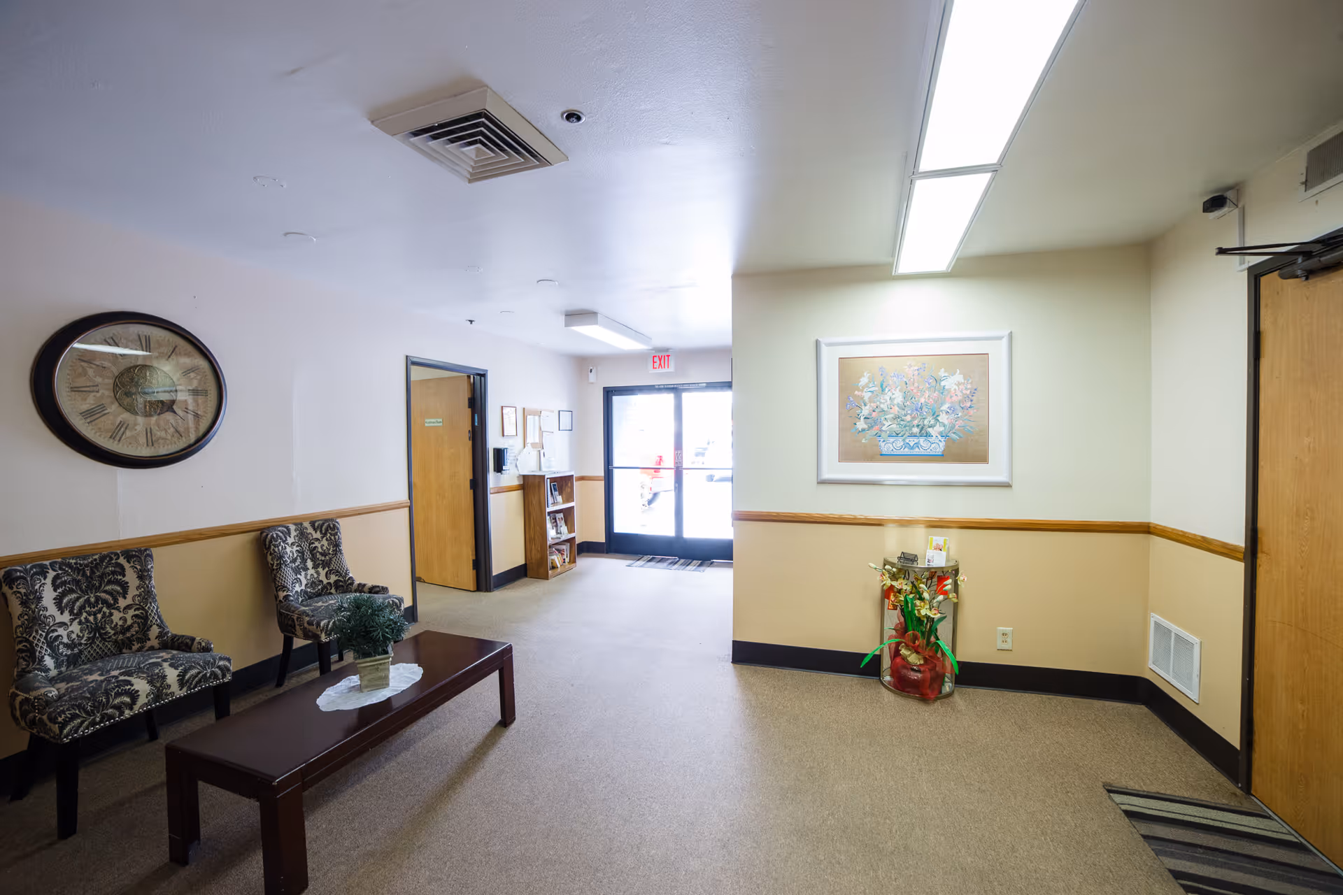 Interior view of a senior living facility lobby area with two patterned chairs, a wooden coffee table with a plant, a large wall clock, a framed floral painting, and a glass door exit in the background.
