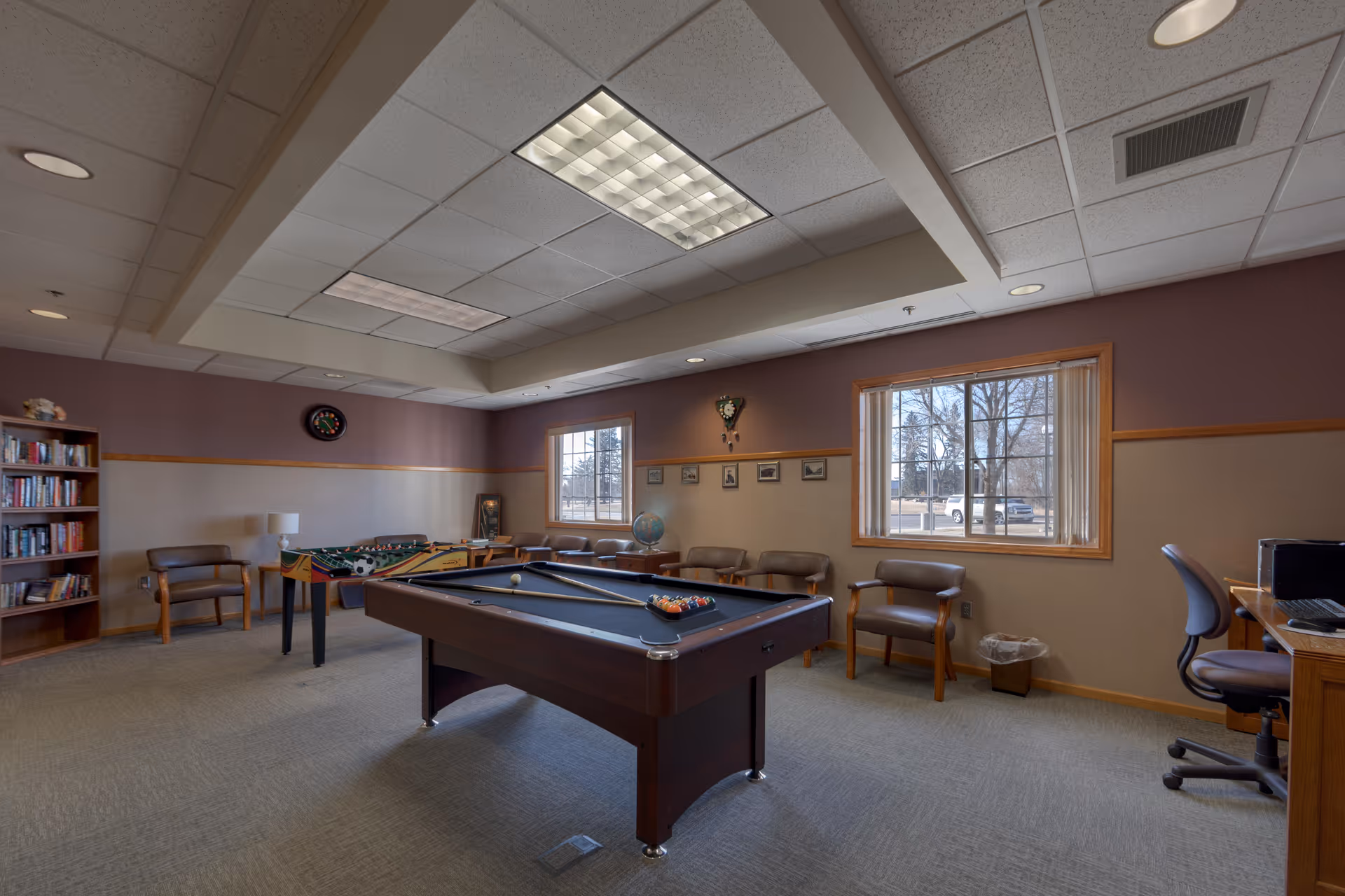 A recreational room with a pool table in the center, a foosball table, several chairs along the walls, a bookshelf filled with books, a dartboard, and a desk with a computer. The room has two windows with a view of trees and a parked vehicle outside. The walls are painted in two tones with a wooden trim, and the ceiling has recessed lighting and fluorescent panels.