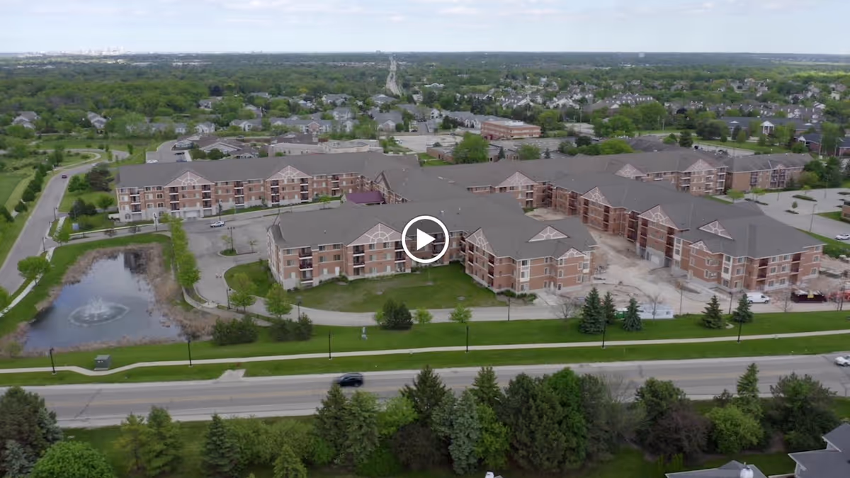 Aerial view of ProHealth Regency Senior Communities New Berlin, showing multiple connected brick buildings with gray roofs, surrounded by green lawns, trees, a pond with a fountain, and nearby roads and residential areas.