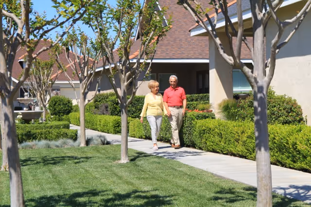 An elderly couple walking along a paved pathway surrounded by neatly trimmed bushes and trees in a sunny outdoor garden area of a residential community.