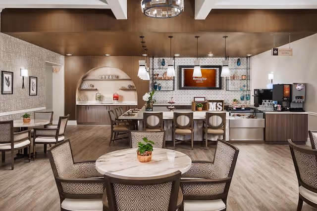A modern dining area in a senior living facility with round and rectangular tables surrounded by cushioned chairs. The room features wood flooring, a decorative wall with shelves holding glassware and decor, pendant lighting, and a coffee and beverage station on the right side. A sign reading 'MorningStar' is displayed on a screen behind the counter.