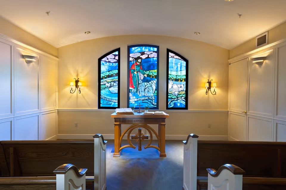 Interior view of a small chapel with wooden pews, a wooden altar with an open Bible and a cross, and three stained glass windows depicting a shepherd with sheep. The walls are light-colored with wall sconces providing warm lighting.