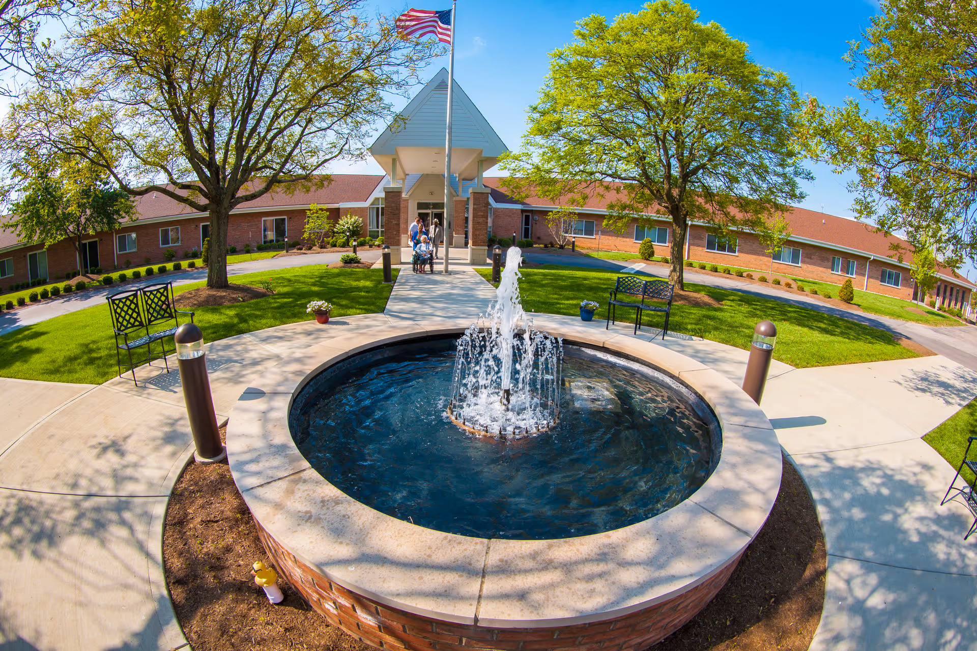 Circular fountain in a landscaped courtyard with benches and trees at the entrance of a single-story brick senior living building.