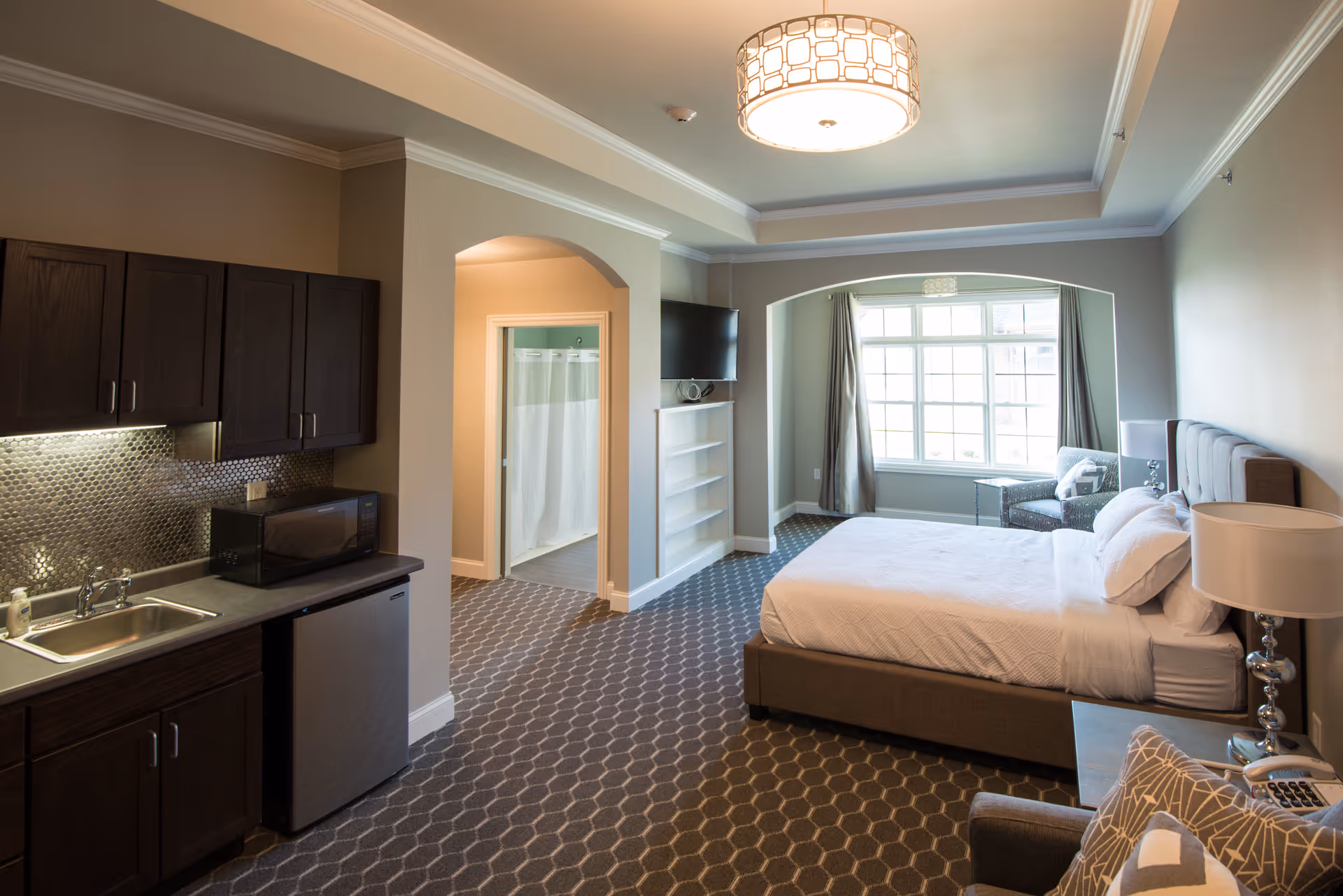 A spacious senior living facility room with a large bed, bedside table with lamp, armchair, and a window with curtains. The room includes a small kitchenette with a sink, microwave, mini fridge, and dark wood cabinets. There is an open doorway leading to a bathroom with a shower curtain visible. The room has patterned carpet flooring and a modern ceiling light fixture.
