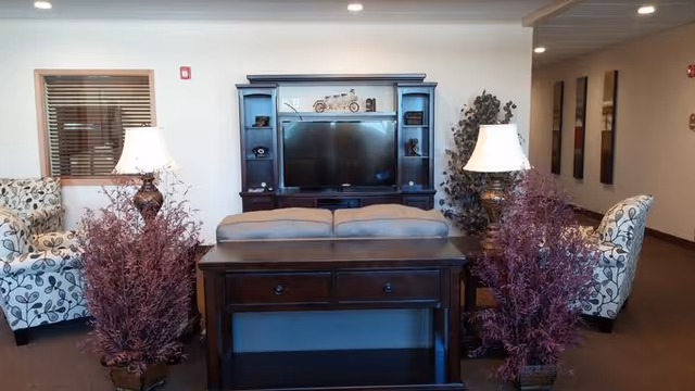 A cozy living room area with a dark wooden entertainment center holding a flat-screen TV and decorative items. In front of the TV is a sofa with a dark wooden console table behind it. On either side of the sofa are two table lamps with white shades and purple decorative plants. Two patterned armchairs are positioned on either side of the room, and there is a window with wooden blinds on the left wall.