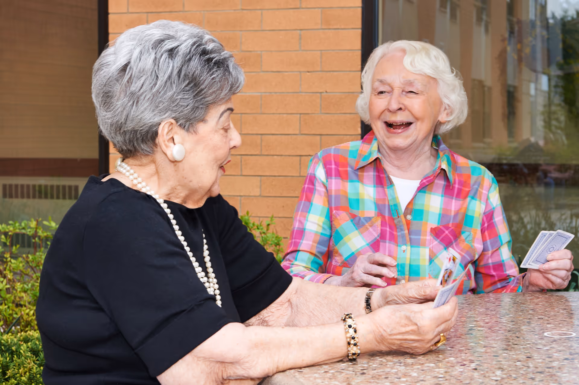 Two elderly women sitting at a table outdoors, playing cards and smiling. One woman is wearing a black top with pearl earrings and necklace, and the other is wearing a colorful plaid shirt.