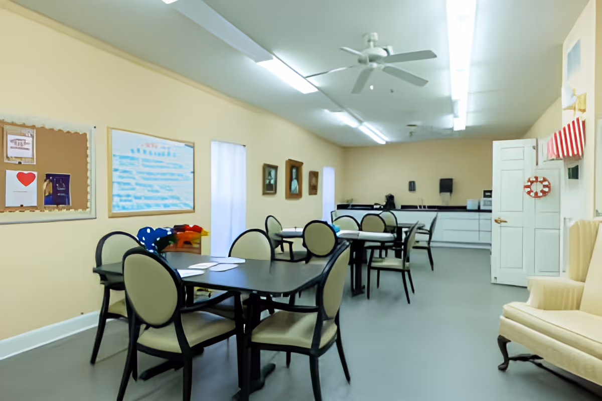 Bright communal activity/dining room with several tables and chairs, bulletin boards on the wall, and a kitchenette in the back.