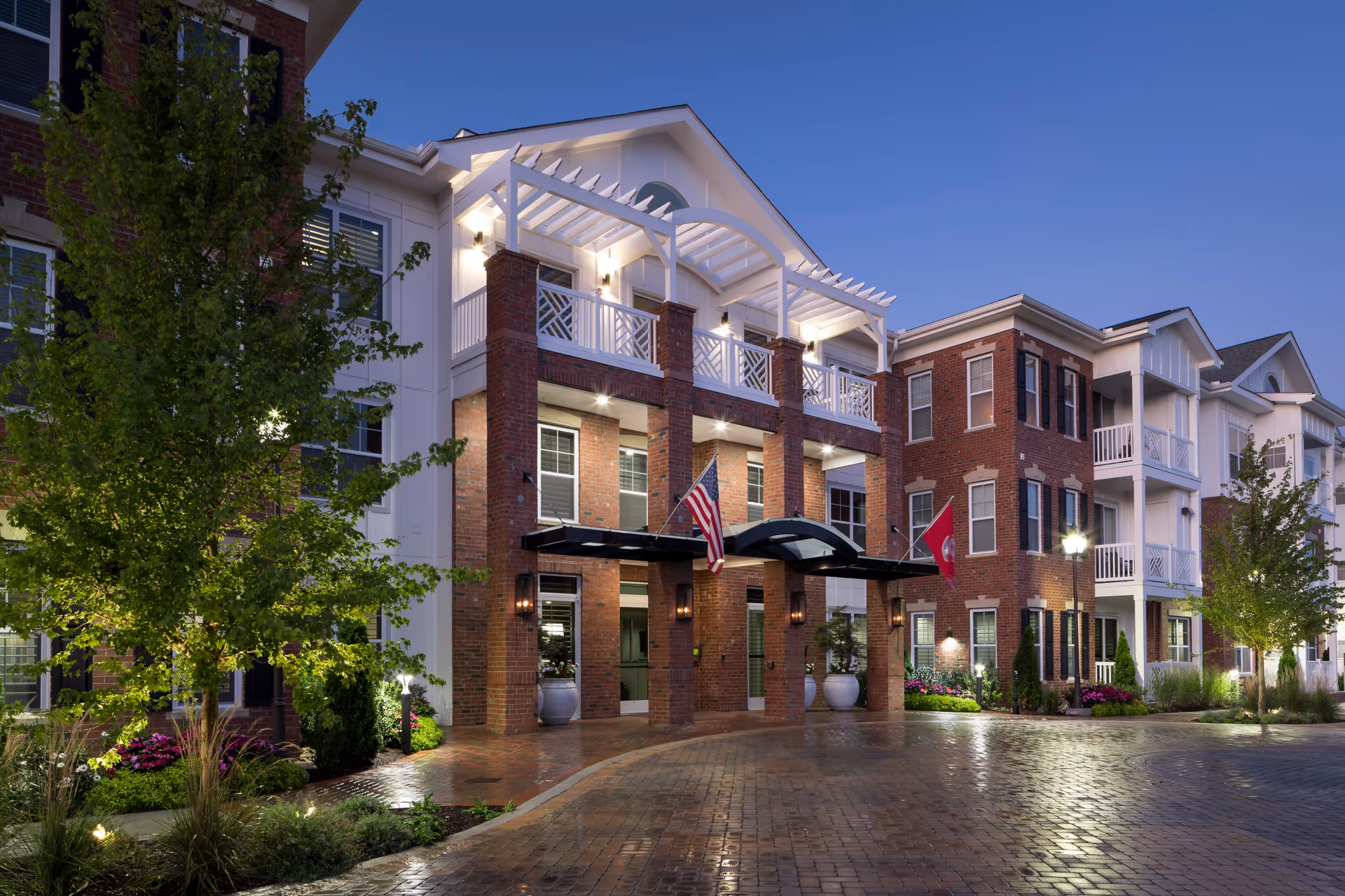 Exterior view of Avenida Cool Springs facility at dusk, showing a well-lit entrance with brick pillars, white railings, and a covered driveway. The building has multiple windows, balconies, and landscaped greenery with trees and flowers along the paved driveway.
