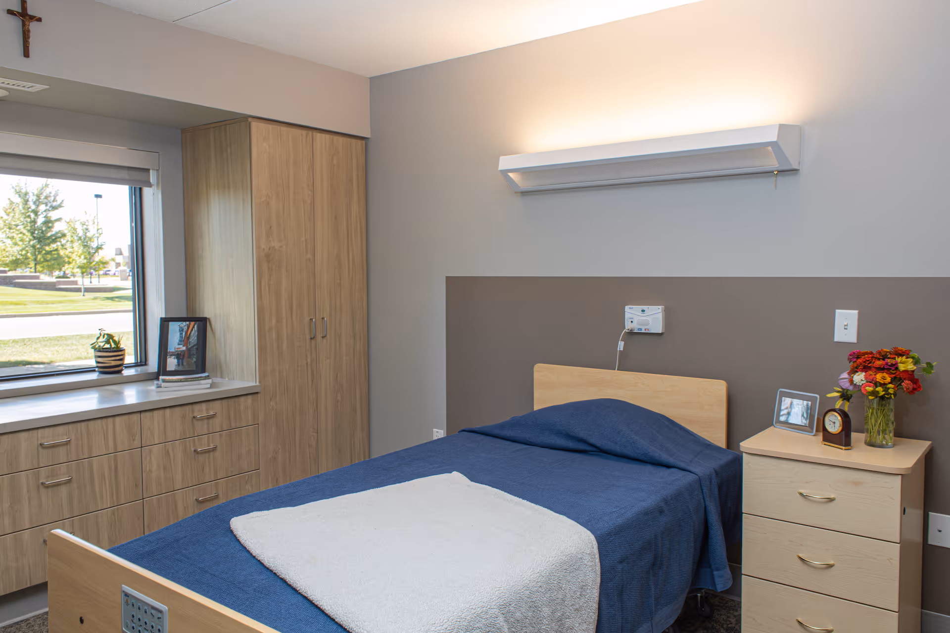 A simple, clean bedroom in a senior living facility with a single bed covered in a blue blanket and a white towel at the foot. There is a wooden nightstand with a clock, a framed photo, and a vase of colorful flowers. A wooden cabinet and drawers are positioned under a window showing an outdoor view with trees and grass. A crucifix hangs on the wall above the window.