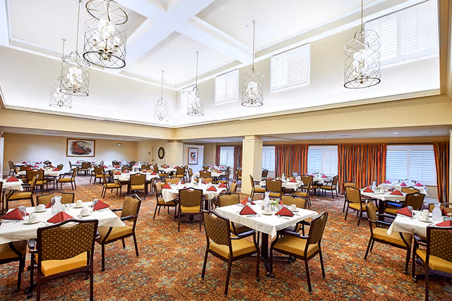 A spacious dining room with multiple tables covered in white tablecloths, each set with red folded napkins, cups, and silverware. The room features patterned carpet, cushioned chairs, large windows with white blinds and orange curtains, and decorative hanging light fixtures from a high ceiling with skylights.