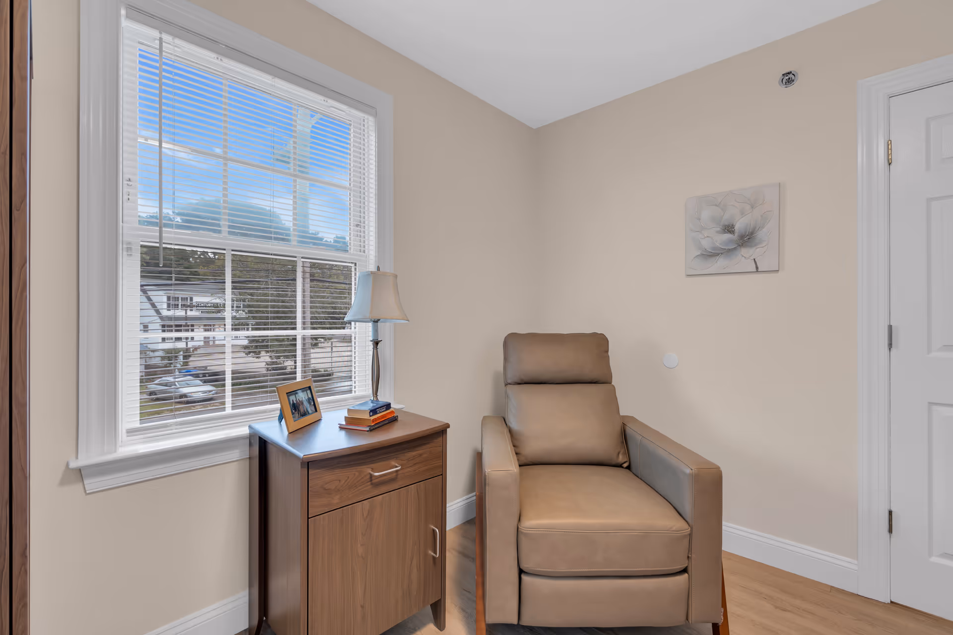 Beige recliner next to a wooden side table with a lamp and framed photo by a window with blinds in a neutral-toned room.