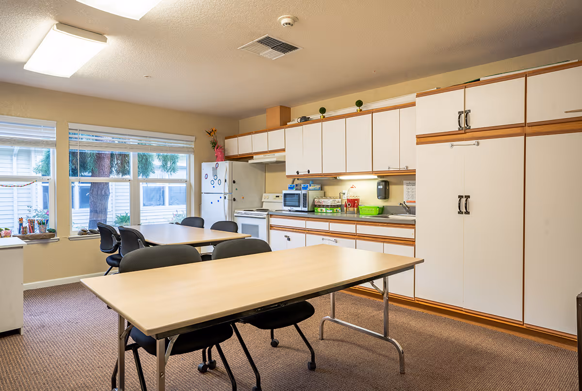 Communal dining room with tables and chairs in front of a kitchenette featuring white cabinets, a refrigerator, microwave and sink.
