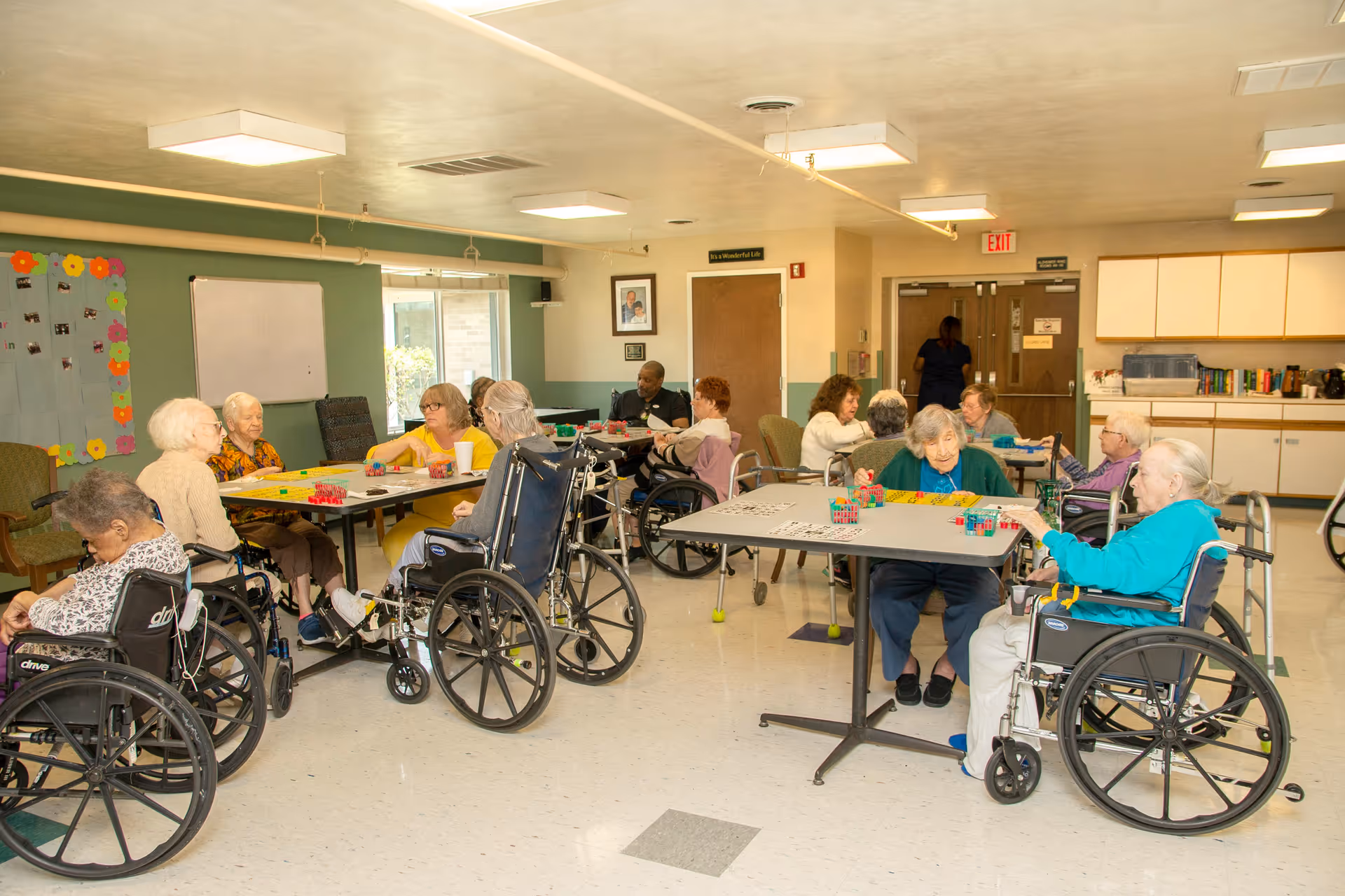 A group of elderly individuals, many in wheelchairs, sitting around tables in a well-lit room playing games and engaging in activities. The room has light-colored walls, a bulletin board with colorful decorations, and cabinets along one wall. A staff member is seen near the exit door in the background.