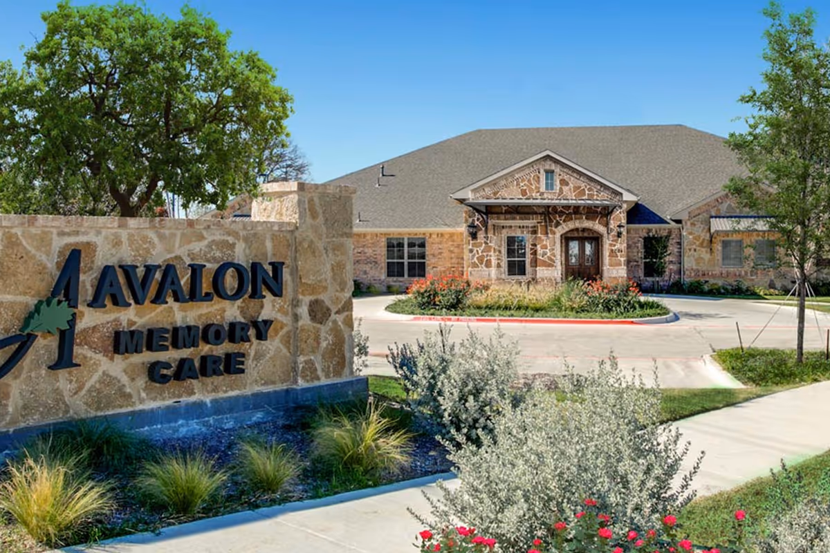 Exterior view of Avalon Memory Care facility with a stone sign in the foreground and a stone building with a peaked roof and wooden double doors in the background, surrounded by landscaping and trees under a clear blue sky.