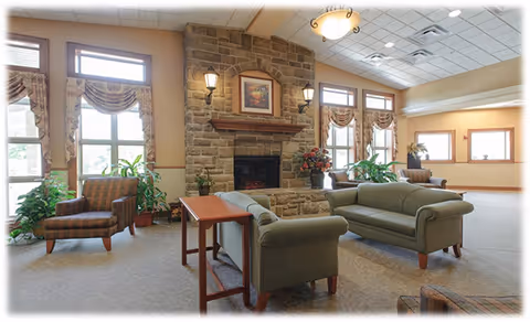 A spacious senior living common area with large windows letting in natural light, featuring a stone fireplace with a framed picture above it, two green sofas, a wooden side table, an armchair, and several potted plants. The room has a high ceiling with recessed lighting and wall sconces on either side of the fireplace.