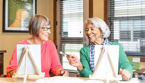 Two elderly women sitting at a table indoors, smiling and engaging in a painting activity with canvases on easels in front of them.