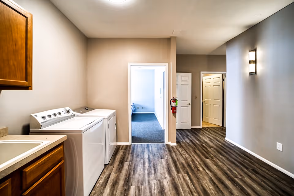 Interior view of a laundry area with a washing machine and dryer on the left side, wooden cabinets above the machines, and a sink countertop. The room has wood-patterned flooring and beige walls. There are two open doorways leading to other rooms, one showing a carpeted bedroom with a bed partially visible, and the other leading to a hallway with closed doors. A fire extinguisher is mounted on the wall near the doorways.