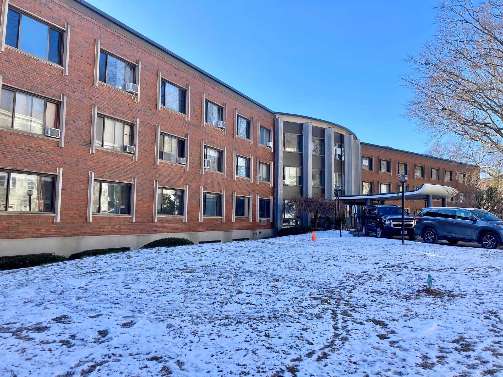 Exterior view of a three-story brick building with multiple windows, some with air conditioning units. The ground is covered with a light layer of snow, and two vehicles are parked near the entrance, which has a covered drop-off area. Leafless trees are visible on the right side under a clear blue sky.