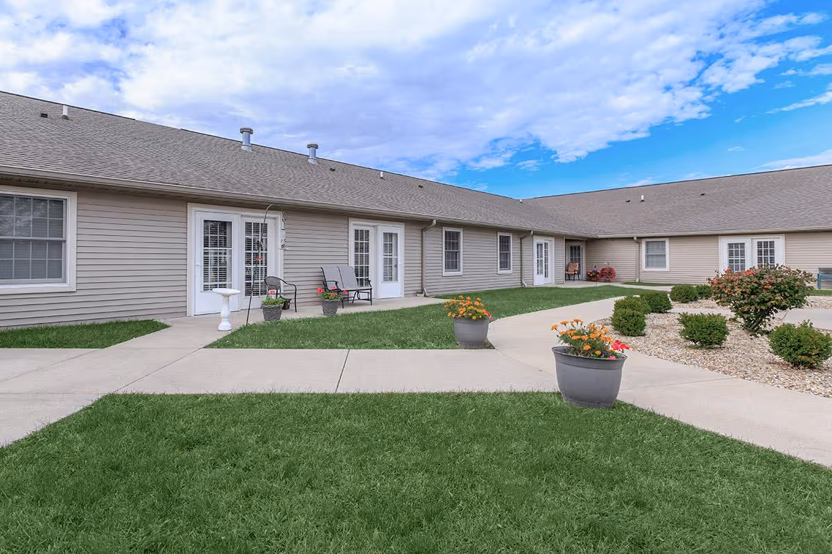 Outdoor courtyard area of a senior living facility with beige siding buildings, multiple white-framed glass doors and windows, concrete walkways, green grass, potted flowers, and small bushes under a partly cloudy blue sky.