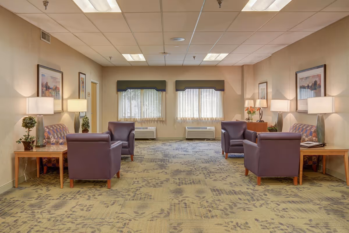 A spacious lounge area in a senior living facility with purple chairs, sofas, tables, lamps, and framed artwork.