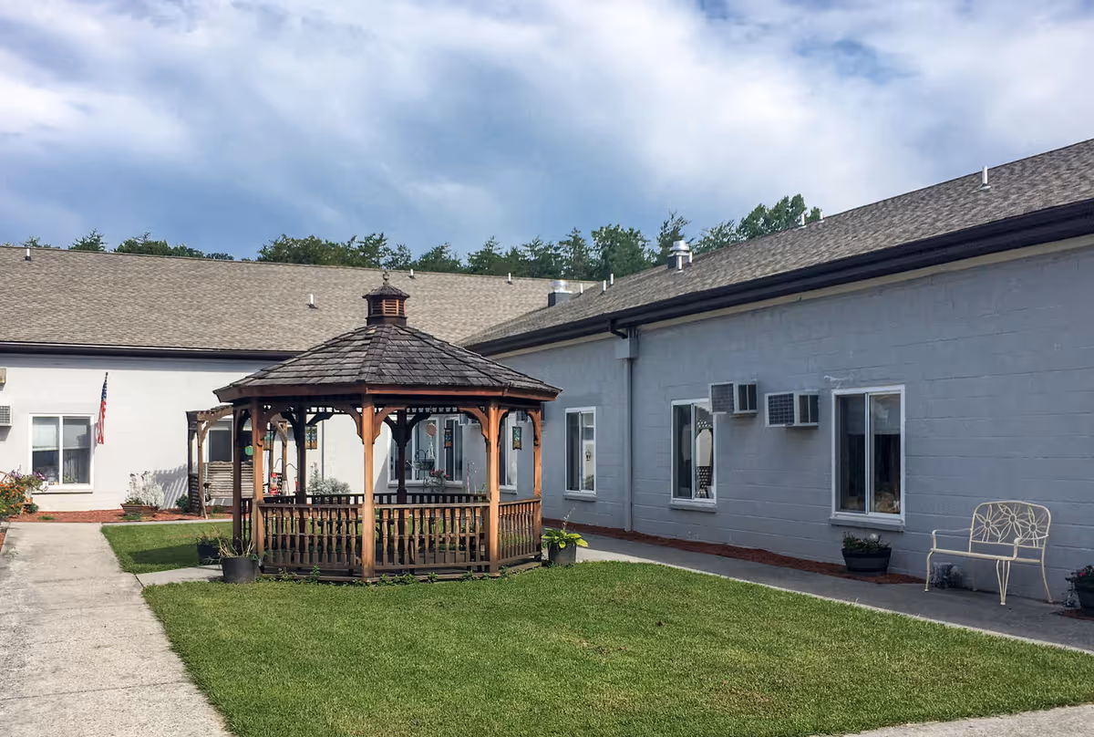 Outdoor courtyard area of Berkeley Springs Healthcare Center featuring a wooden gazebo in the center, surrounded by a well-maintained lawn and concrete walkways. The building exterior is light gray with several windows and air conditioning units. There is a white metal bench and some potted plants near the building walls. The sky is partly cloudy.