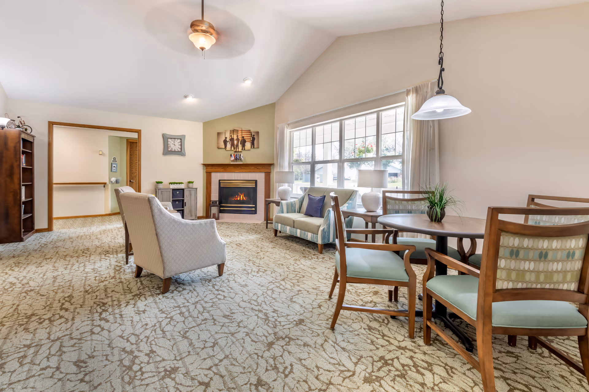 Bright common living room with seating, a fireplace, large window, and a dining table in a senior living facility.