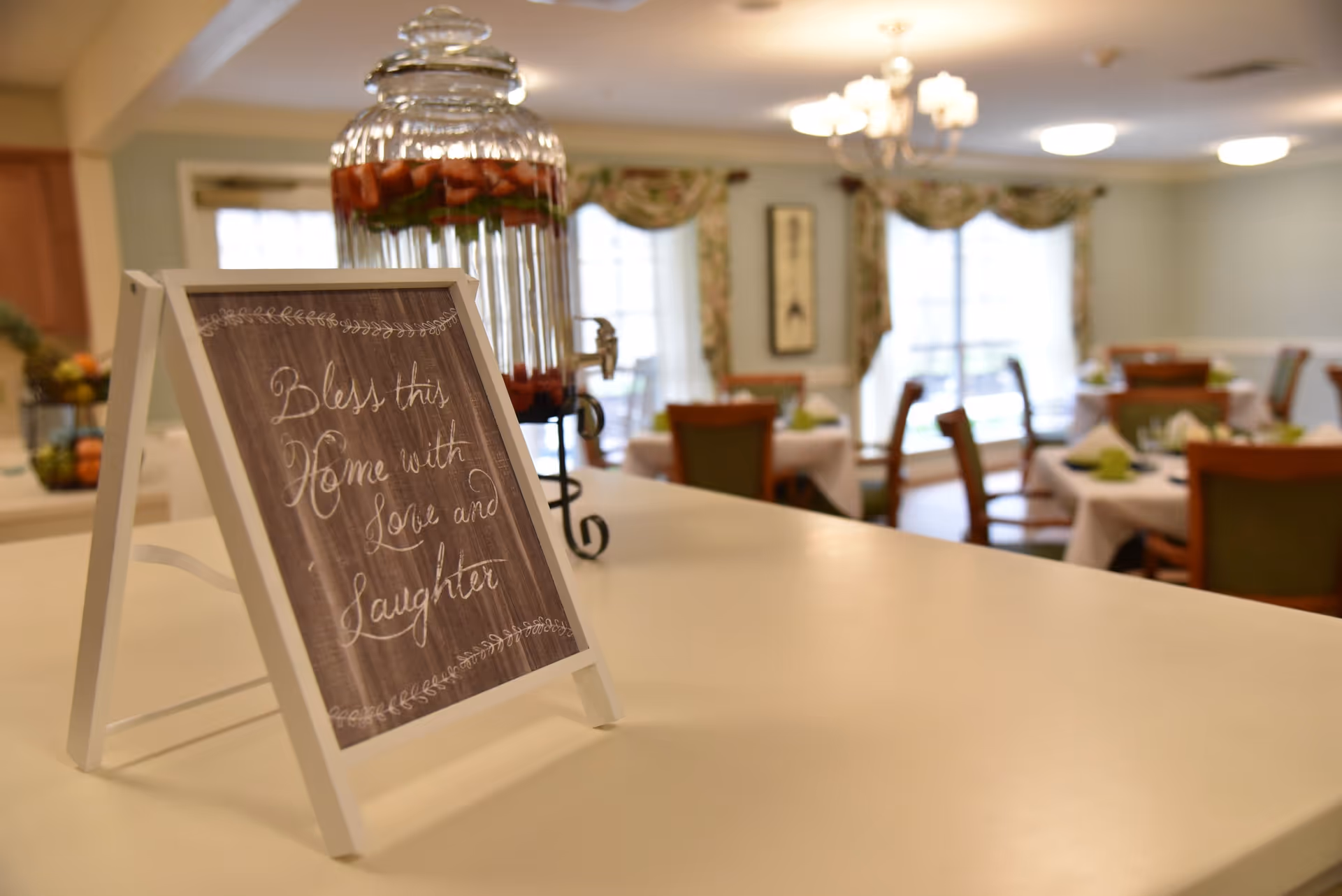 A dining area in a senior living facility with tables set with white tablecloths and green napkins. In the foreground, there is a small framed sign on a counter that reads 'Bless this Home with Love and Laughter' and a glass beverage dispenser filled with water and fruit. The room has large windows with floral curtains and a chandelier overhead.