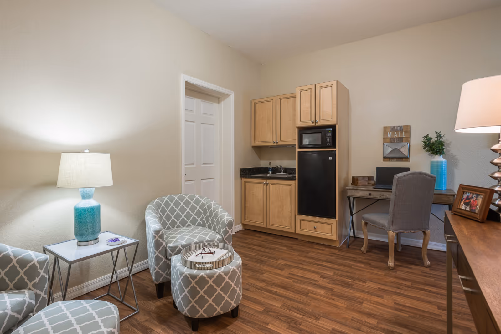 Small living area with two patterned armchairs and an ottoman, side table with a lamp, a kitchenette with fridge and microwave, and a desk.