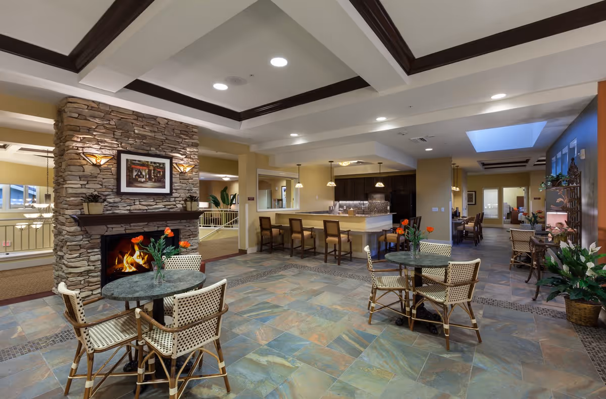 A spacious and well-lit common area featuring a stone fireplace with a fire burning, two round tables each surrounded by four wicker chairs, a kitchen counter with bar stools, and a dining area in the background. The floor is tiled with a mix of earth tones, and the ceiling has recessed lighting and decorative beams.