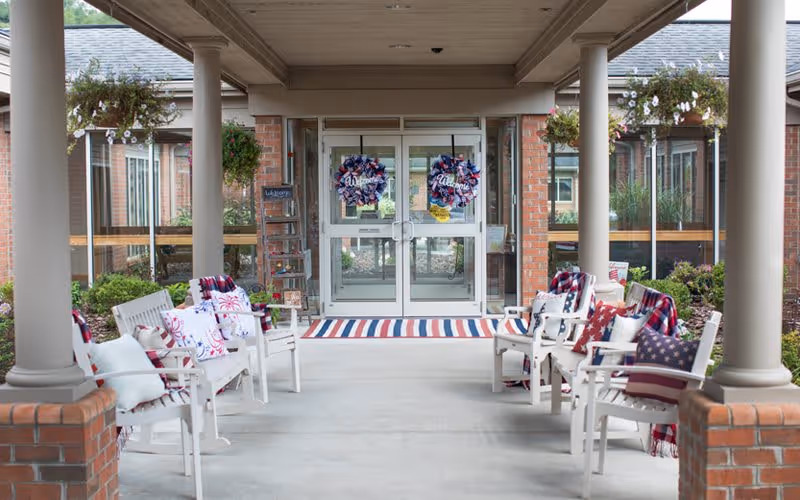 Covered outdoor seating area at the entrance of a building with white chairs adorned with patriotic-themed pillows and blankets. The entrance has double glass doors decorated with wreaths, and there are hanging flower baskets on both sides.