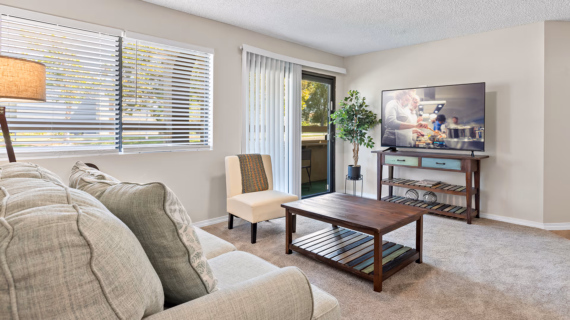A bright living room with a beige sofa and a single cream-colored chair with a decorative pillow. There is a wooden coffee table in the center and a wooden TV stand holding a flat-screen television displaying a cooking show. A potted plant is placed next to the TV stand. The room has large windows with blinds and a sliding glass door leading outside. The carpet is light beige, and the walls are painted a soft neutral color.