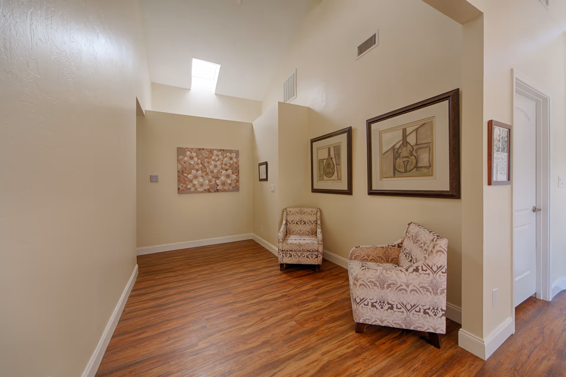 Small sitting area with two patterned armchairs, framed artwork on the walls, wood flooring, and a skylight.