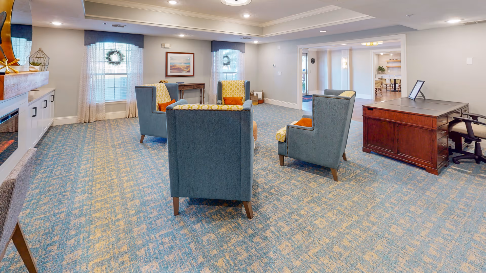Bright communal lounge with upholstered armchairs arranged around a seating area, patterned carpet, windows with curtains, and a wooden desk.