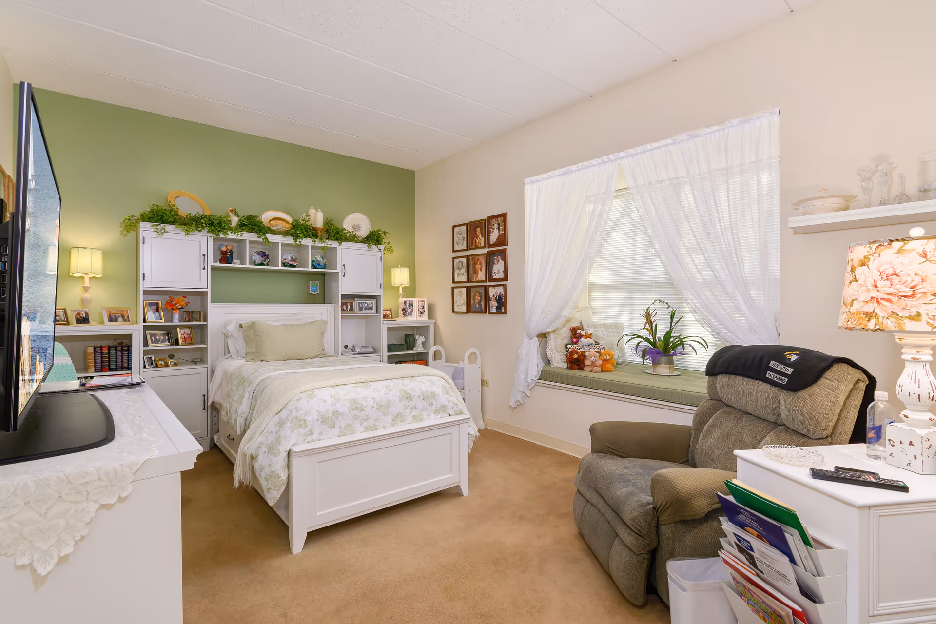 A cozy bedroom with a white bed featuring floral bedding against a green accent wall. The bed is flanked by white shelves and cabinets decorated with plants, photos, and ornaments. A large window with sheer white curtains and a cushioned window seat with stuffed animals and a potted plant is on the right. A comfortable recliner chair and a white side table with a floral lamp and magazines are in the foreground. A flat-screen TV is on a white dresser on the left side of the room.