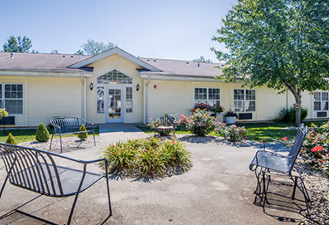 Outdoor patio area with metal benches and chairs arranged around a small garden bed with plants and flowers, in front of a single-story building with white siding and multiple windows under a clear blue sky.