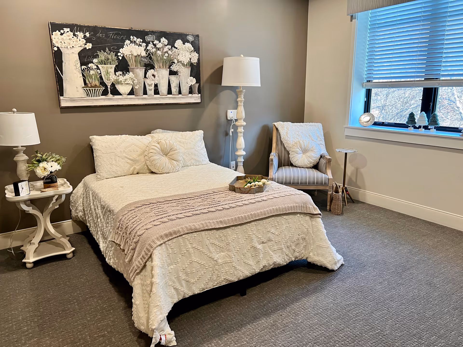 A cozy bedroom with a bed covered in a textured white bedspread and a light pink knitted throw blanket. The bed has two round decorative pillows. To the left of the bed is a white side table with a lamp, a small clock, and a flower arrangement. Above the bed hangs a large painting of white flowers in various vases. To the right of the bed is a tall white floor lamp and a striped armchair with a white throw blanket and a round pillow. A window with blue blinds lets in natural light, and small decorative items are placed on the windowsill.