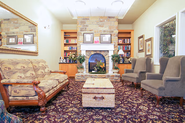 A cozy living room with a patterned carpet, a floral upholstered wooden sofa on the left, two gray armchairs on the right, and a wicker coffee table in the center. The back wall features a stone fireplace with a white mantle, flanked by built-in wooden bookshelves filled with books and decorative items. There are framed pictures on the walls and a large mirror above the sofa reflecting part of the room.