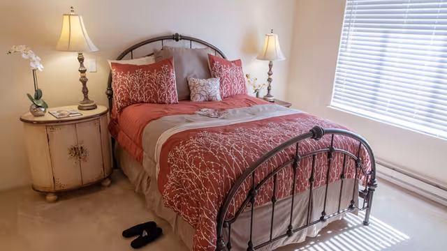 A cozy bedroom with a metal framed bed dressed in red and beige bedding with floral patterns. There are two matching bedside tables, each with a lamp and decorative items. A pair of black slippers is placed on the carpeted floor near the bed. Sunlight streams in through a window with white blinds.
