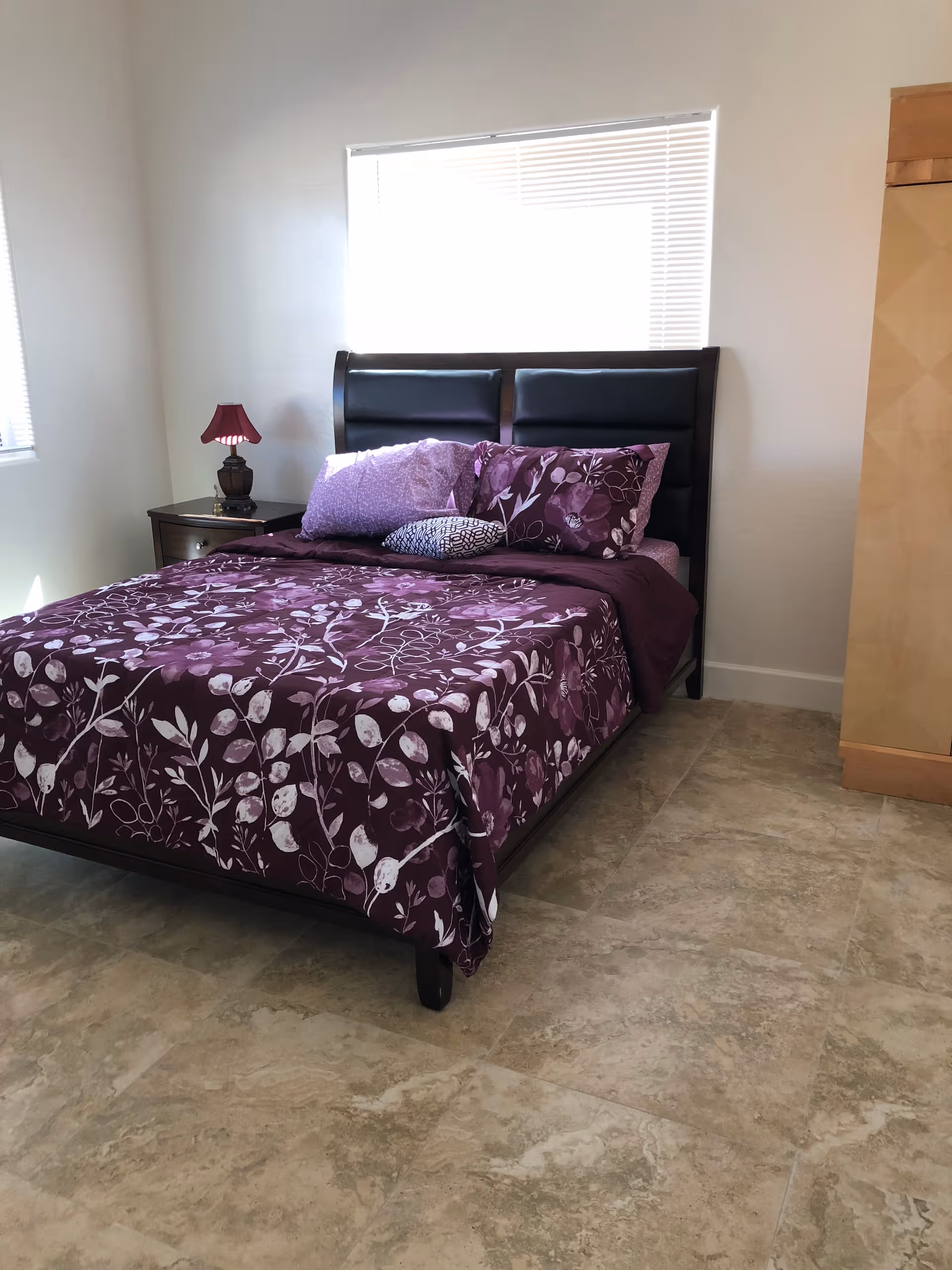 A bedroom with a bed featuring a dark wooden frame and a purple floral bedspread with matching pillows. There is a small nightstand with a red lamp on the left side of the bed, a window with blinds behind the bed, and a light-colored wooden wardrobe on the right side. The floor is tiled with beige tiles.