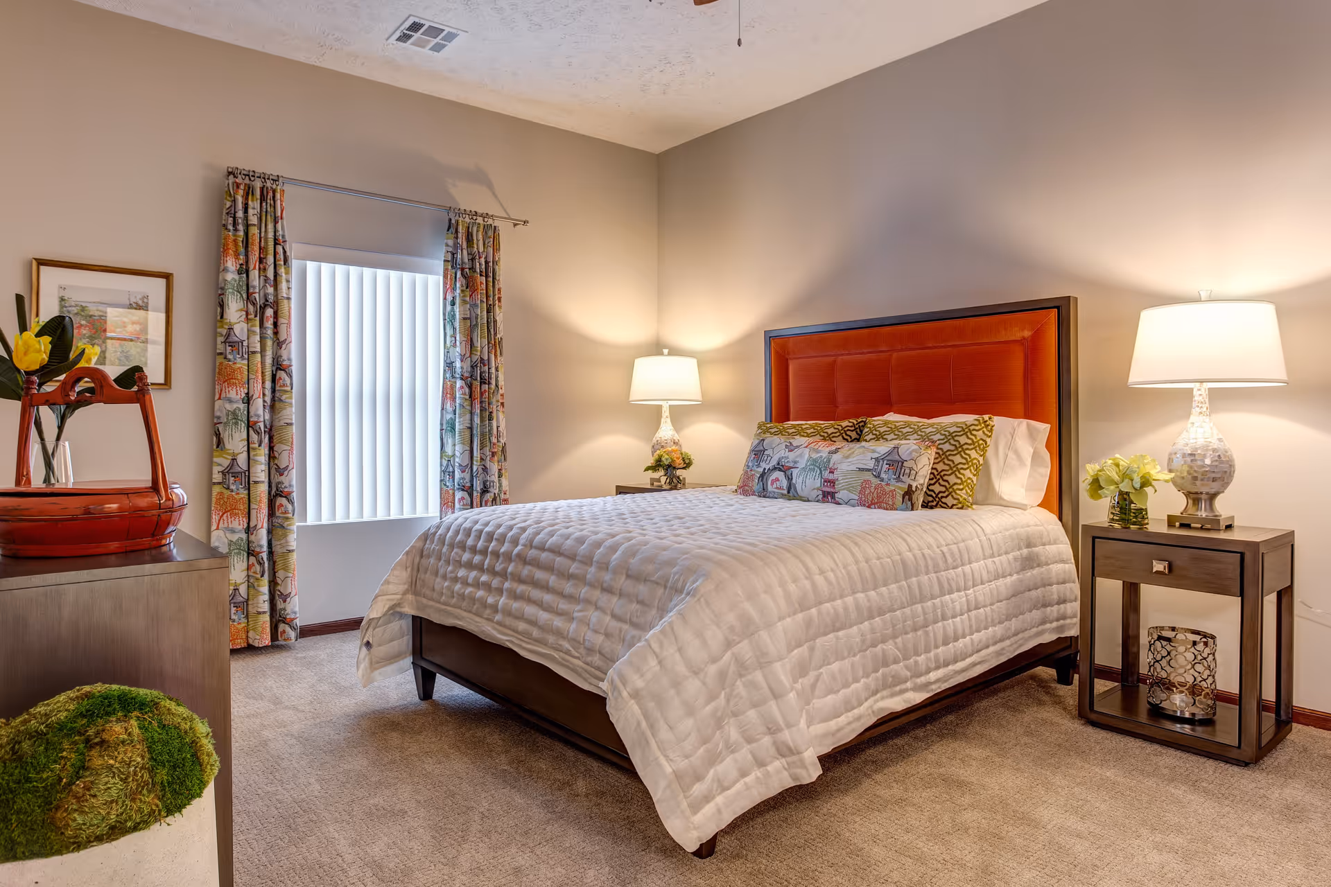 A cozy bedroom in Hudson Meadows Retirement Community featuring a bed with a red upholstered headboard, white quilted bedding, and decorative pillows. There are two bedside tables with lamps and flowers, a window with patterned curtains, a dresser with a red basket and a yellow flower, and a green moss-like decorative item on the floor.