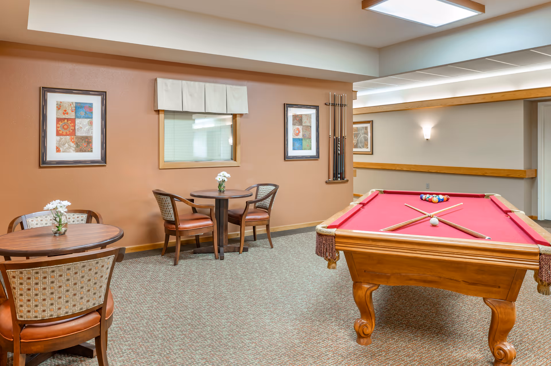 A recreational room with a red felt pool table and two crossed pool cues on it. There are two small round tables with chairs around them, each table decorated with a small vase of white flowers. The walls are painted in a warm beige color and adorned with framed artwork. The room has carpeted flooring and soft overhead lighting.