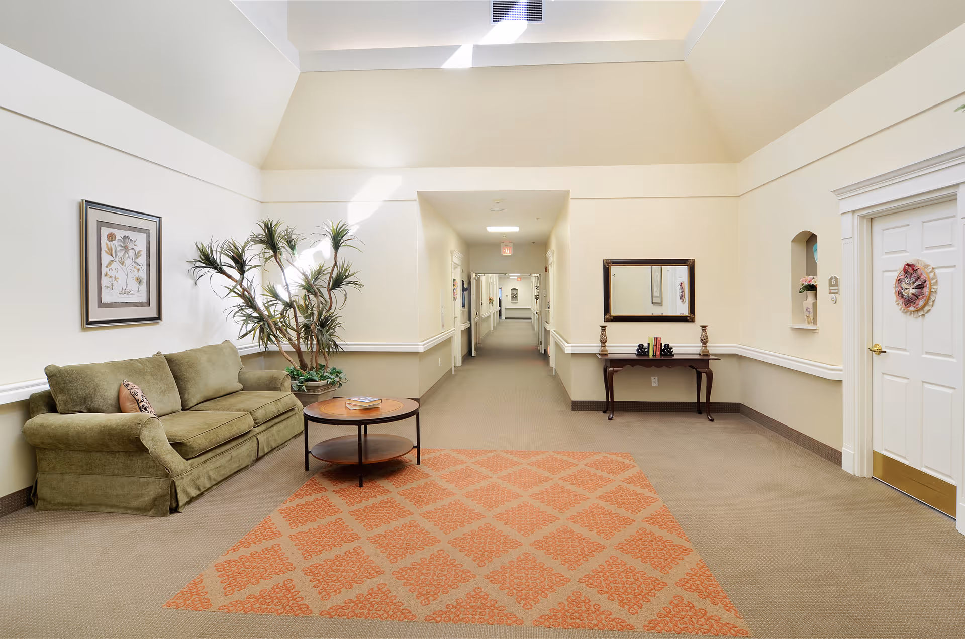 A bright hallway in a senior living facility with beige walls and carpet. On the left side, there is a green sofa with a small round wooden coffee table in front of it, and a tall potted plant next to the sofa. A framed botanical print hangs above the sofa. On the right side, there is a wooden console table with decorative items and a mirror above it. A white door with a decorative wreath is also visible on the right. The hallway extends into the distance with doors and artwork along the walls.
