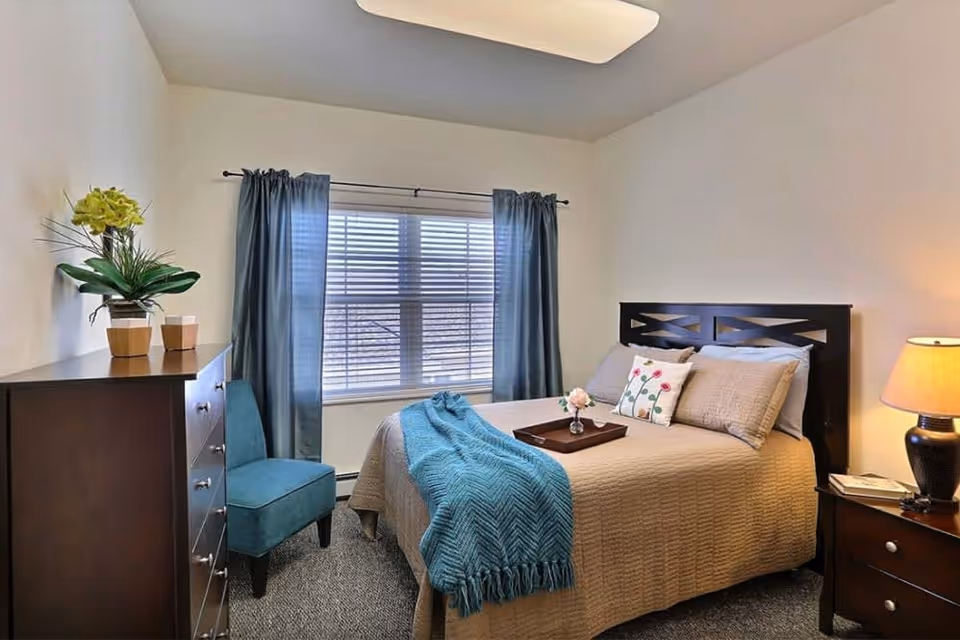Furnished bedroom with a made bed topped by a teal throw and decorative pillows, a teal chair, wooden dresser, window with curtains, and a bedside lamp.