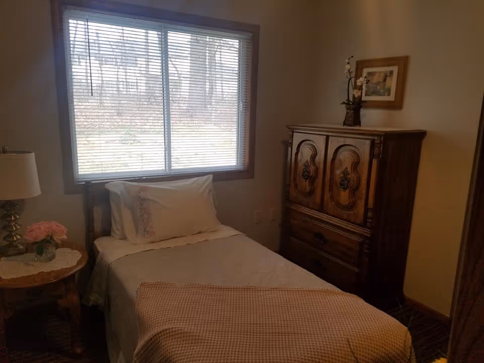 A small bedroom with a single bed covered in a white bedspread and a beige blanket at the foot. There is a pillow with floral embroidery on the bed. Next to the bed is a wooden nightstand with a lamp and a vase of pink flowers. A large window with blinds lets in natural light. Across from the bed is a wooden dresser with ornate carvings, topped with a small plant and a framed picture on the wall above it.