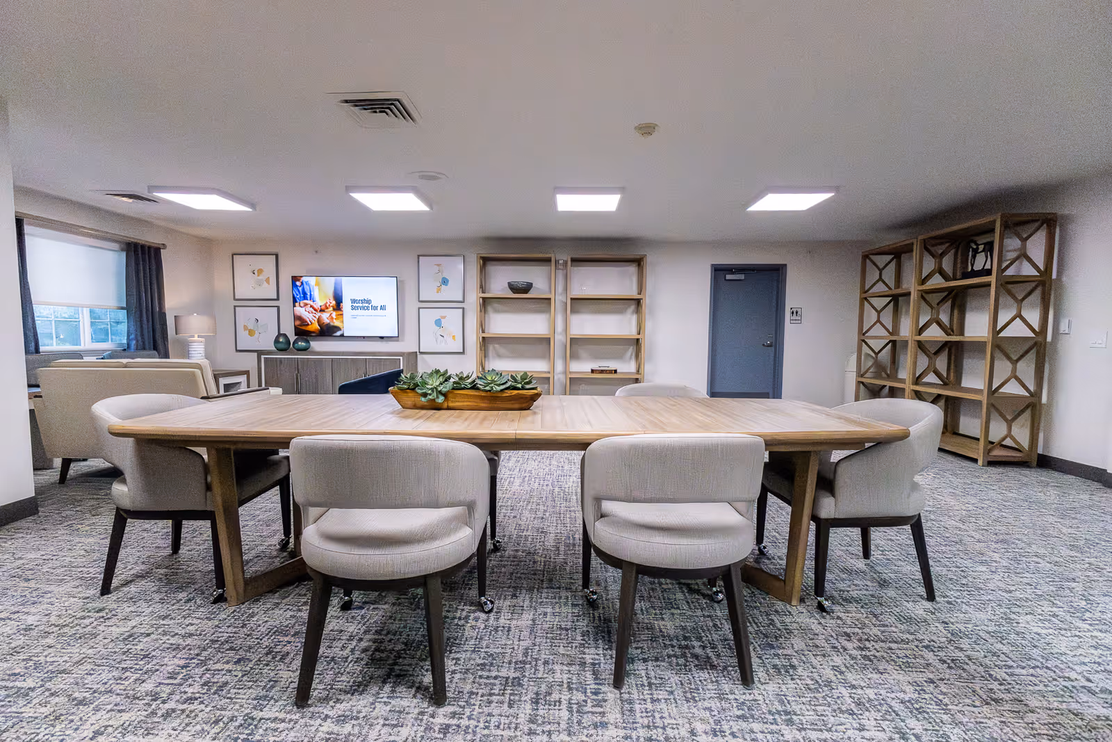A communal dining/activity room with a long wooden table surrounded by upholstered chairs, shelving and a TV on the far wall.