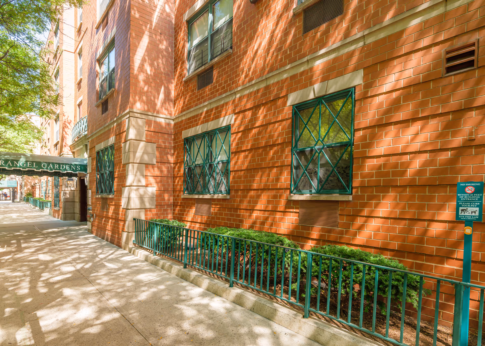 Exterior view of a brick building with green window grilles and a green awning that reads 'RANGEL GARDENS'. There are small bushes behind a green metal fence along the sidewalk, and a no smoking sign is visible near the building.