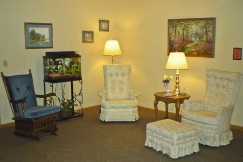 A cozy sitting area in an assisted living facility with three upholstered chairs, including a rocking chair, a small wooden side table with a lamp and flowers, and a fish tank on a stand. The walls are decorated with framed paintings and the room is softly lit by two lamps.